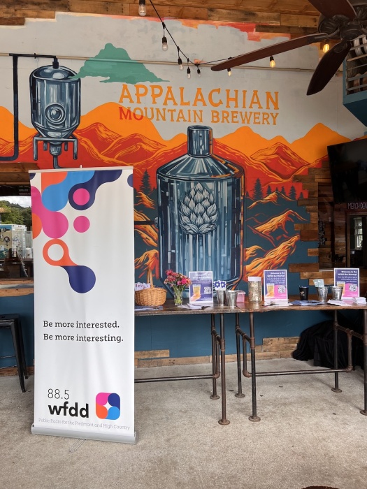 A table with flyers and information on supporting WFDD stands in front of a mural at Appalachian Mountain Brewery.