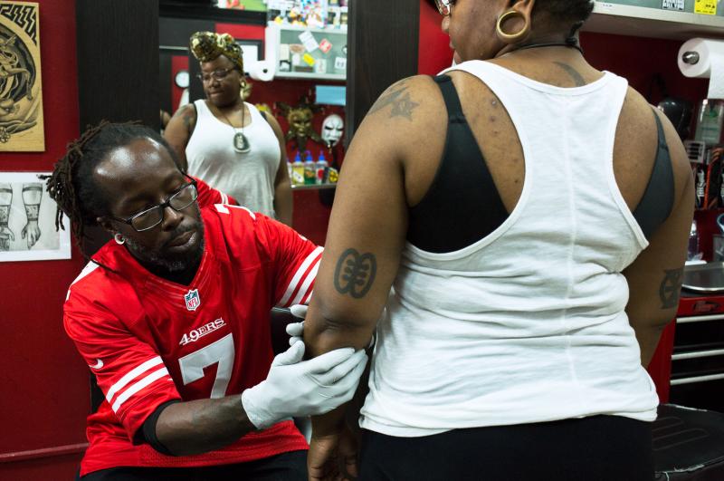 Christopher Mensah, owner and tattoo artist at the Pinz-N-Needlez tattoo shop in Washington, D.C., creates the outline for Oshun Afrique's 35th tattoo. Christopher Mensah, owner and tattoo artist at the Pinz-N-Needlez tattoo shop in Washington, D.C., creates the outline for Oshun Afrique's 35th tattoo.