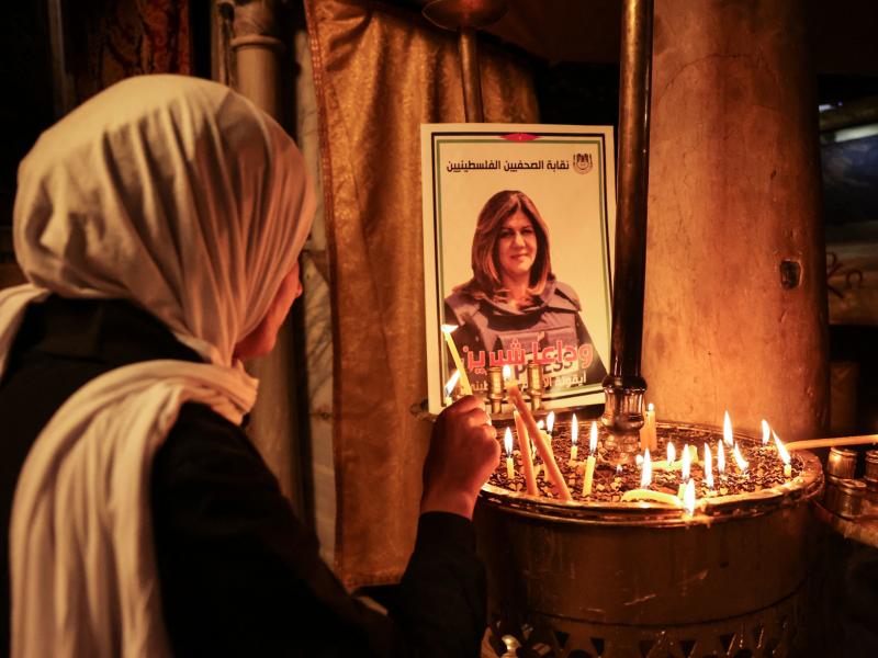 A woman lights a candle in front of a poster depicting Shireen Abu Akleh. A woman lights a candle in front of a poster depicting Shireen Abu Akleh.