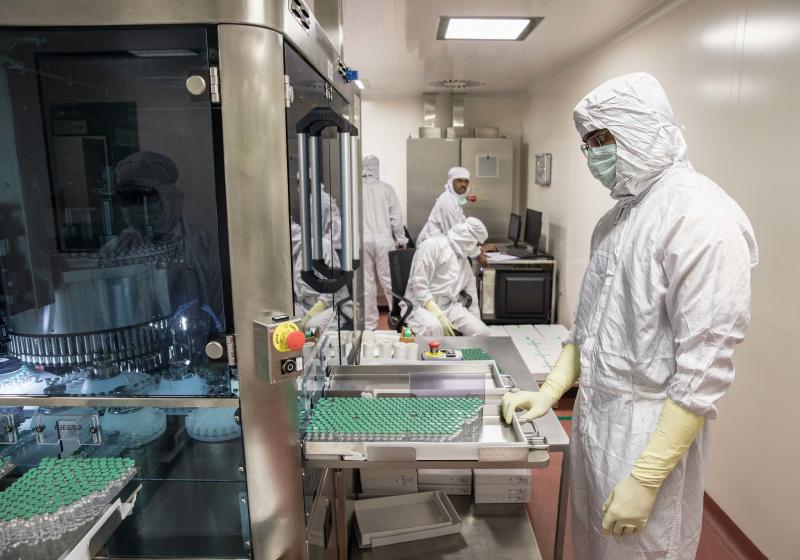 A technician waits to collect vials containing vaccine after they pass through a machine that checks for bottling and vaccine substance deficiencies. A technician waits to collect vials containing vaccine after they pass through a machine that checks for bottling and vaccine substance deficiencies.