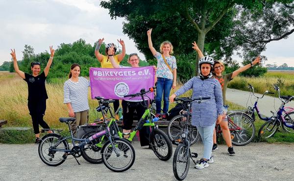 Volunteers and trainees with the group Bikeygees at a park in Berlin in July. The organization teaches refugee women in Germany how to ride bikes. Trainee Shapol Bakir-Rasoul, a refugee from Iraq, holds up a Bikeygees sign with founder Annette Krüger, ri Volunteers and trainees with the group Bikeygees at a park in Berlin in July. The organization teaches refugee women in Germany how to ride bikes. Trainee Shapol Bakir-Rasoul, a refugee from Iraq, holds up a Bikeygees sign with founder Annette Krüger, ri