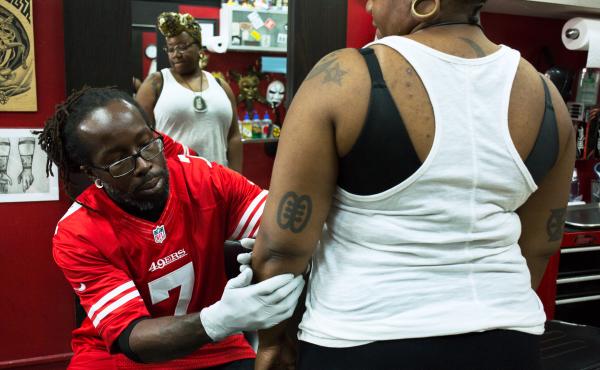 Christopher Mensah, owner and tattoo artist at the Pinz-N-Needlez tattoo shop in Washington, D.C., creates the outline for Oshun Afrique's 35th tattoo. Christopher Mensah, owner and tattoo artist at the Pinz-N-Needlez tattoo shop in Washington, D.C., creates the outline for Oshun Afrique's 35th tattoo.
