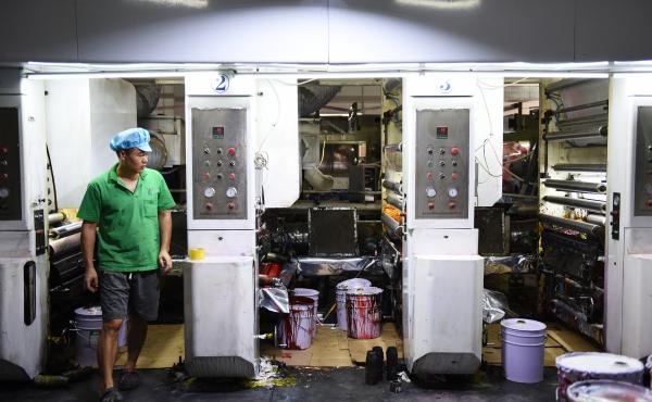 Workers in green polo shirts and blue caps monitor machines making plastic products at the Dongguan Fangjie Printing and Packaging Company. Workers in green polo shirts and blue caps monitor machines making plastic products at the Dongguan Fangjie Printing and Packaging Company.