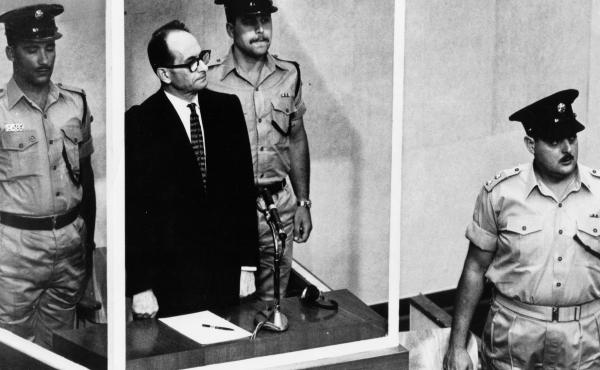 Adolf Eichmann, standing in a glass booth and flanked by guards, in the Jerusalem courtroom during his trial in 1961 for war crimes committed during World War II. Adolf Eichmann, standing in a glass booth and flanked by guards, in the Jerusalem courtroom during his trial in 1961 for war crimes committed during World War II.